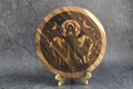 Wooden circular artwork of Hecate with engraved design on a stand against a gray background