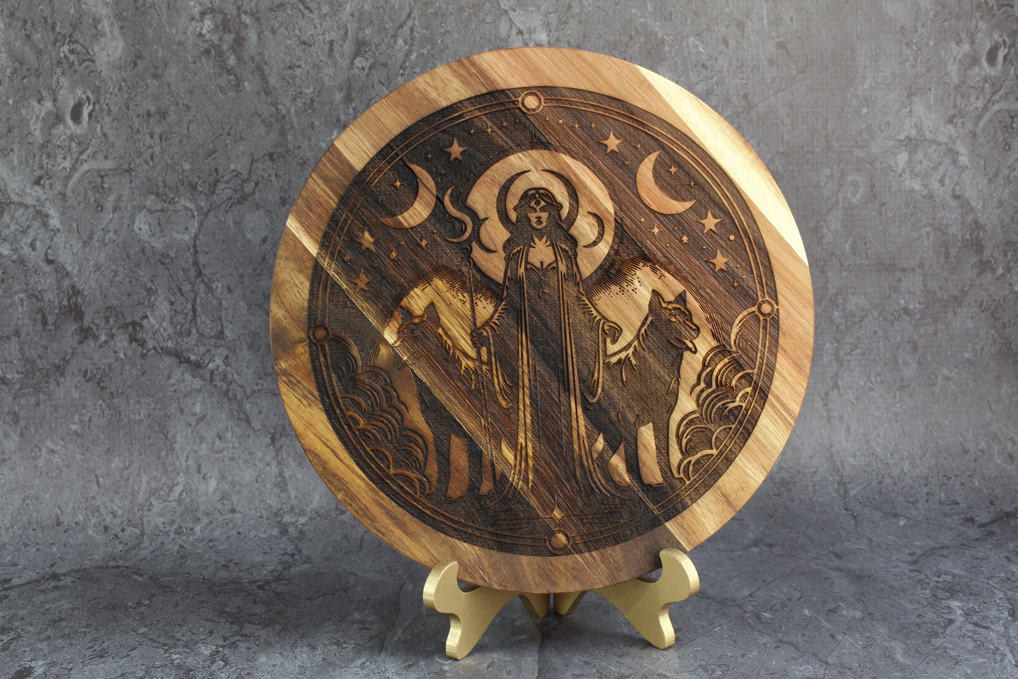 Wooden circular artwork of Hecate with engraved design on a stand against a gray background