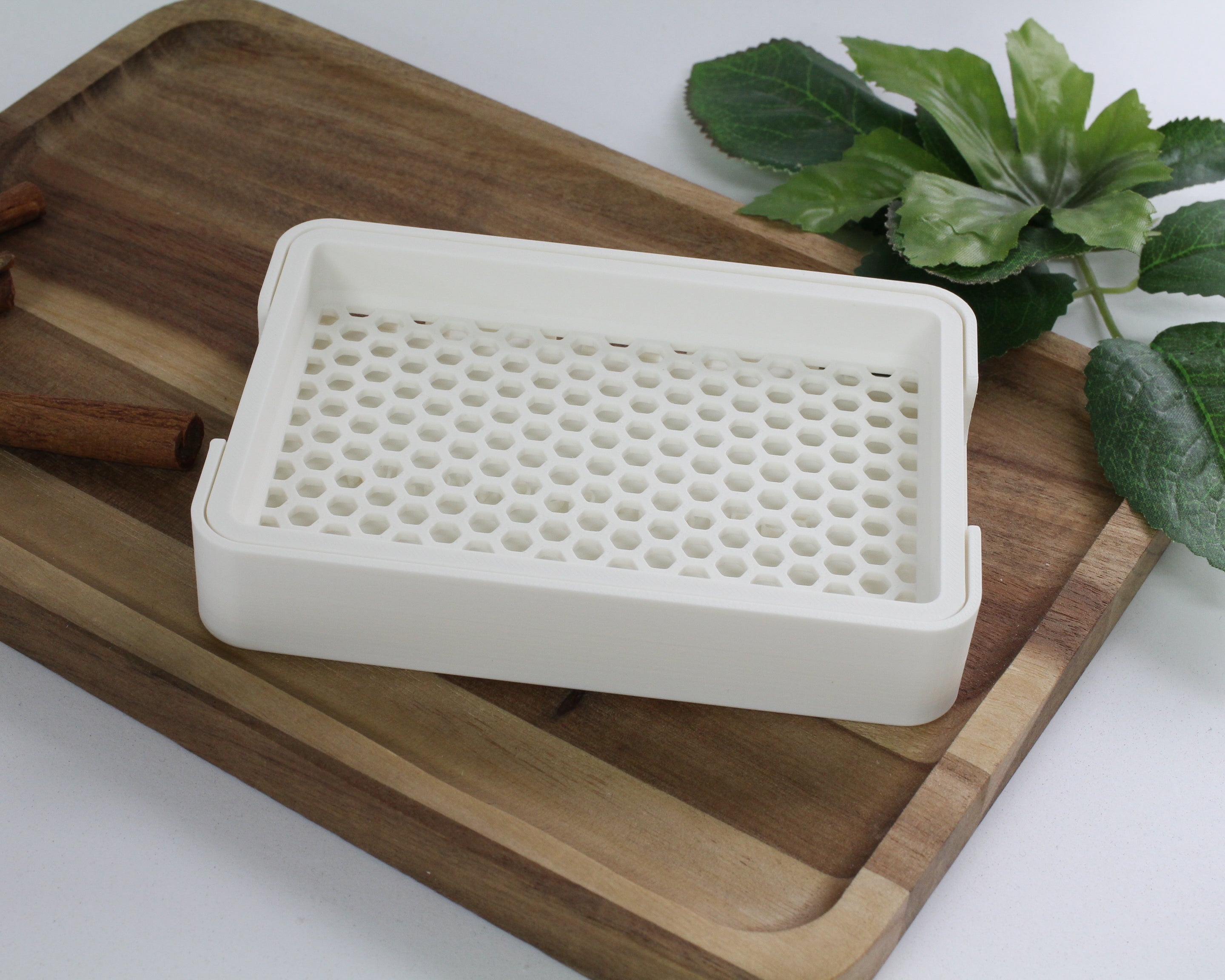 White 3D printed soap dish on a wooden cutting board with cinnamon sticks and green leaves in the background