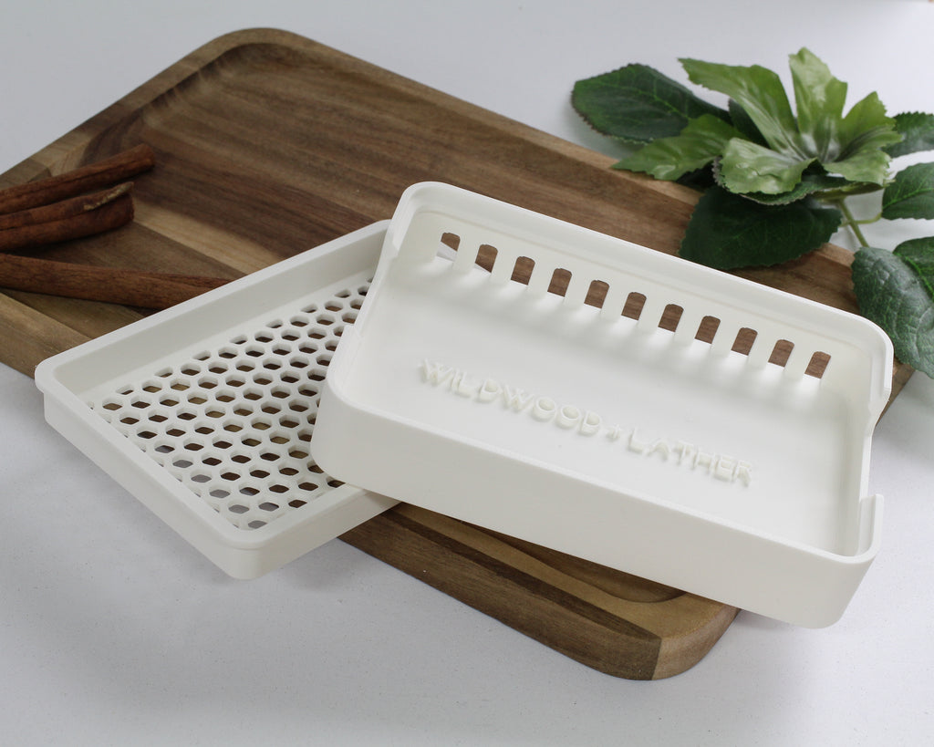 White soap dish with 'Wildwood & Lather' branding on a wooden tray with cinnamon sticks and a plant in the background.