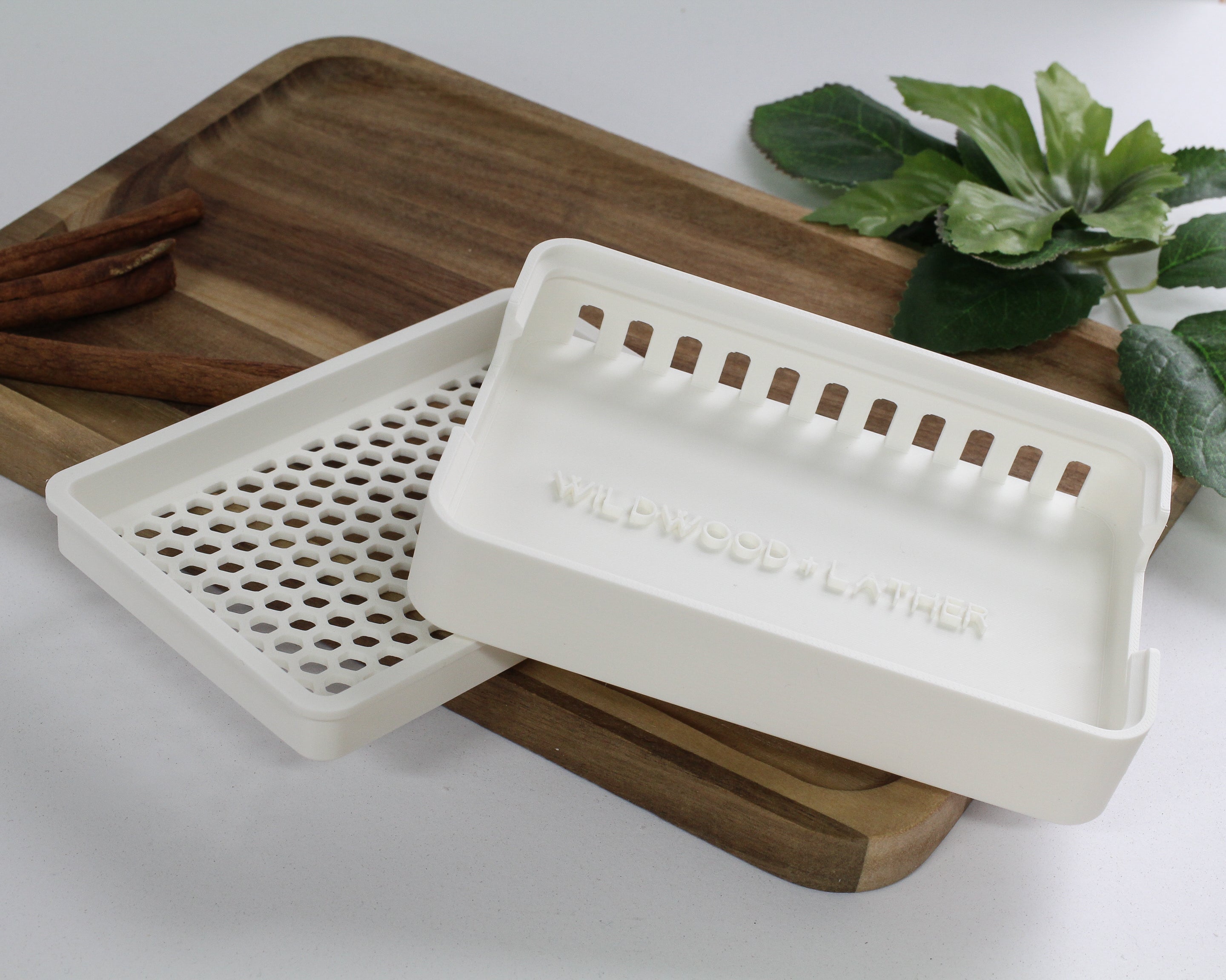 White soap dish with 'Wildwood & Lather' branding on a wooden tray with cinnamon sticks and a plant in the background.