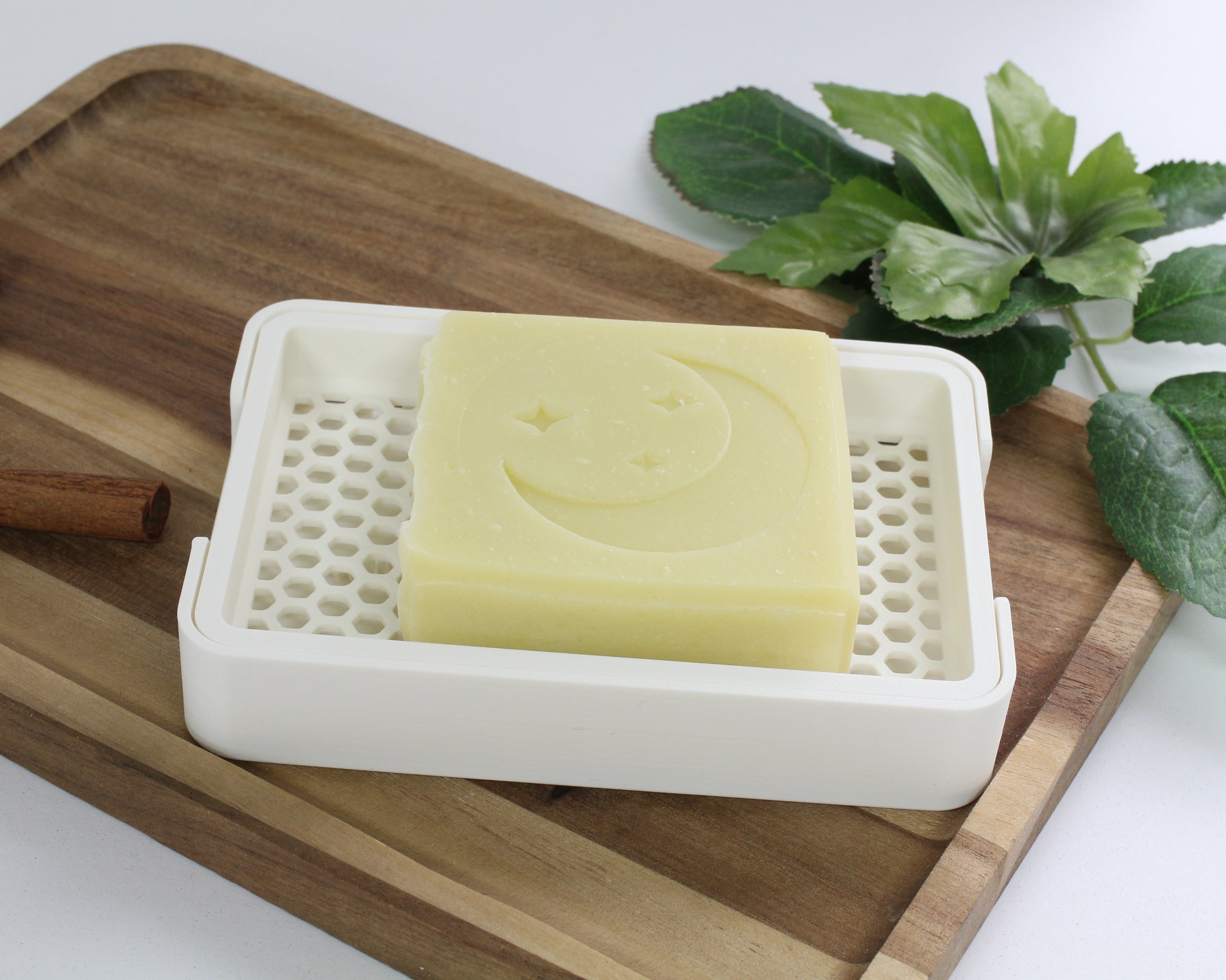 Bar of soap in a white dish on a wooden tray with a cinnamon stick and green leaves in the background.