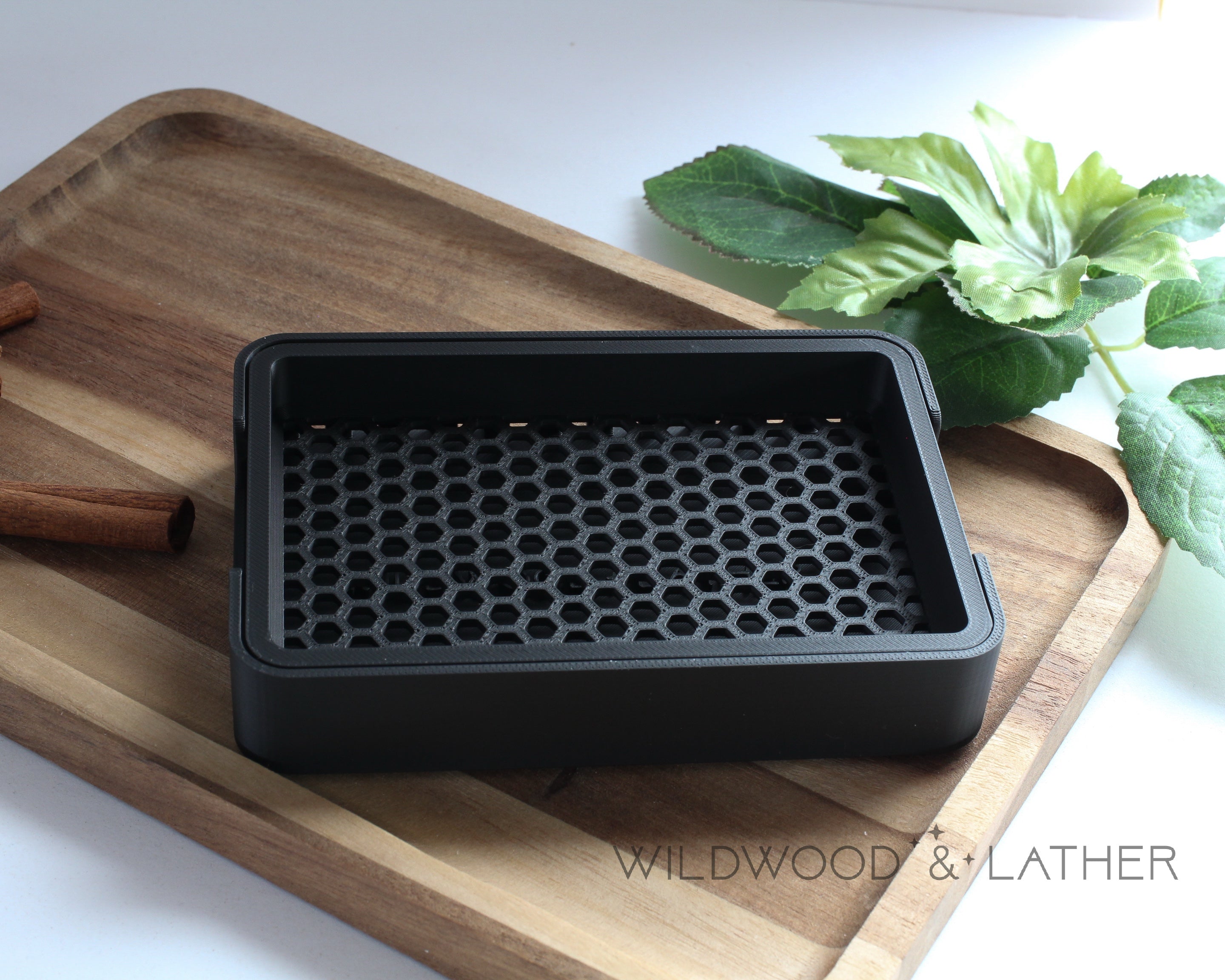 Black 3D printed soap dish on a wooden cutting board with cinnamon sticks and green leaves in the background